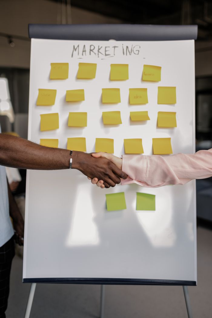 home-img Colleagues shaking hands in front of a marketing board during a business meeting.